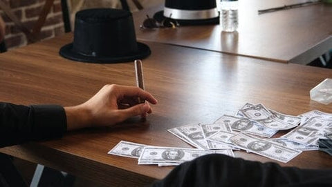 hand holding a cigar on table with money scattered on it